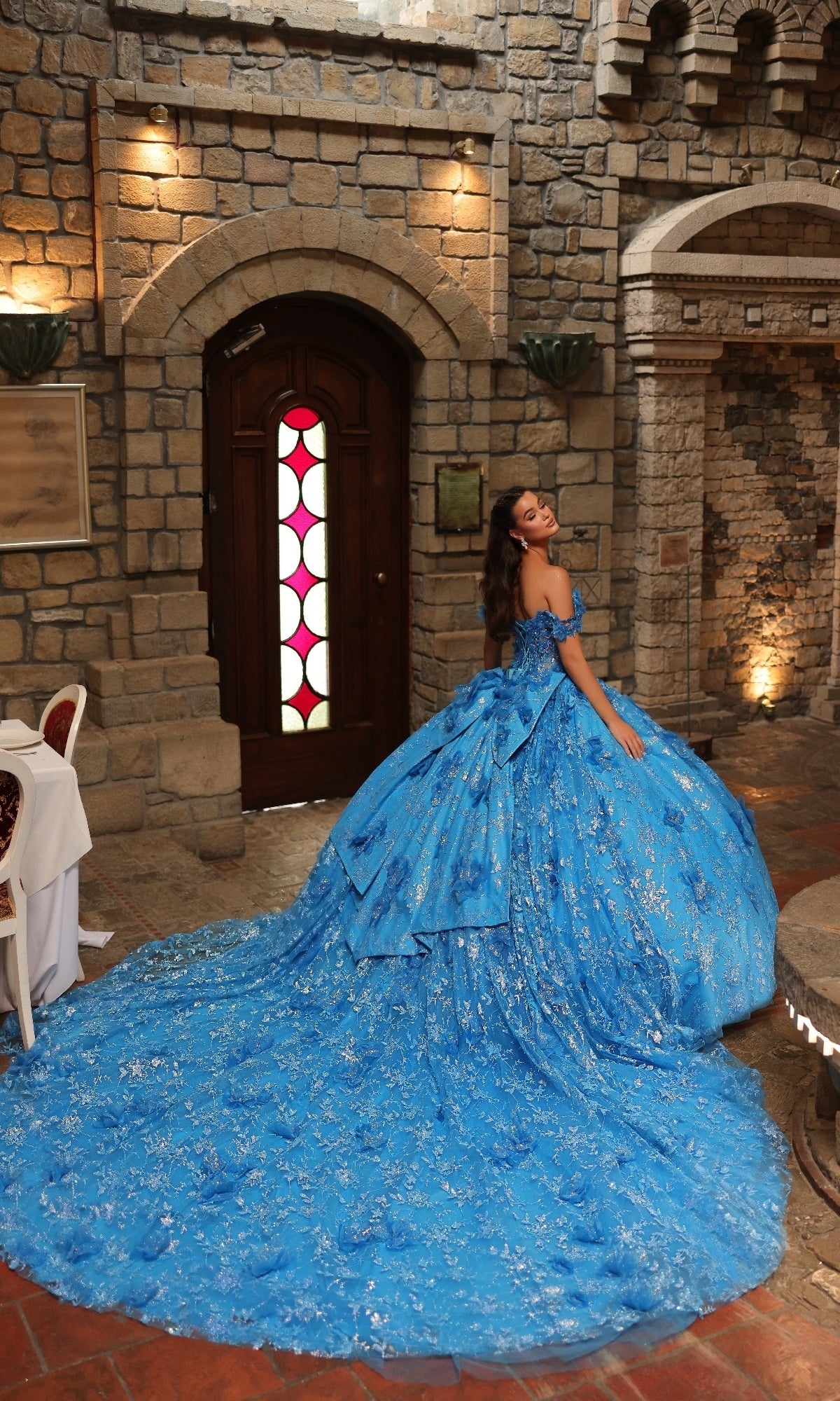 A woman wearing the Off-Shoulder Quinceanera Dress: Amarra 54243, a bright blue ball gown with 3D floral appliques and a long train, smiles over her shoulder in front of a wooden door in a stone-walled room.